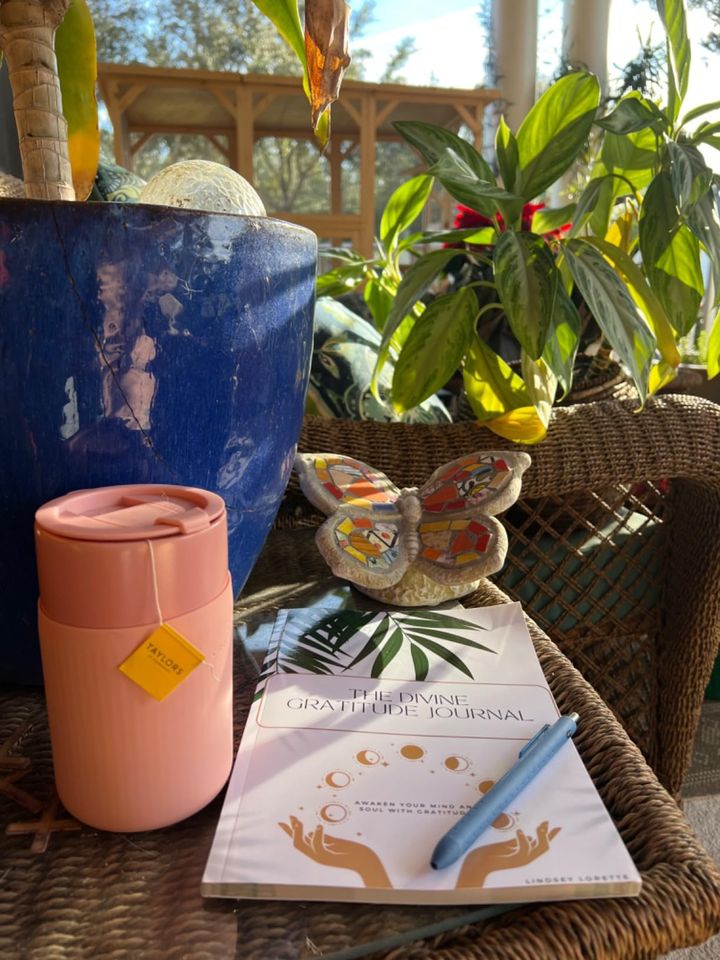 The Divine Gratitude Journal on a sunny wicker table beside a pink tumbler and a mosaic butterfly.