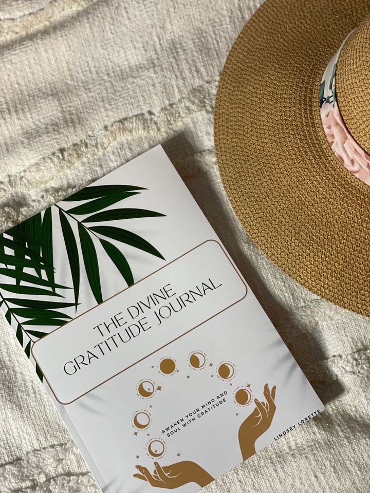 The Divine Gratitude Journal lying on a cream knit blanket beside a straw hat.