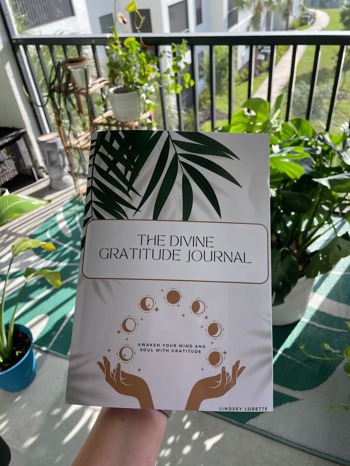 The Divine Gratitude Journal held up on a balcony among green plants.